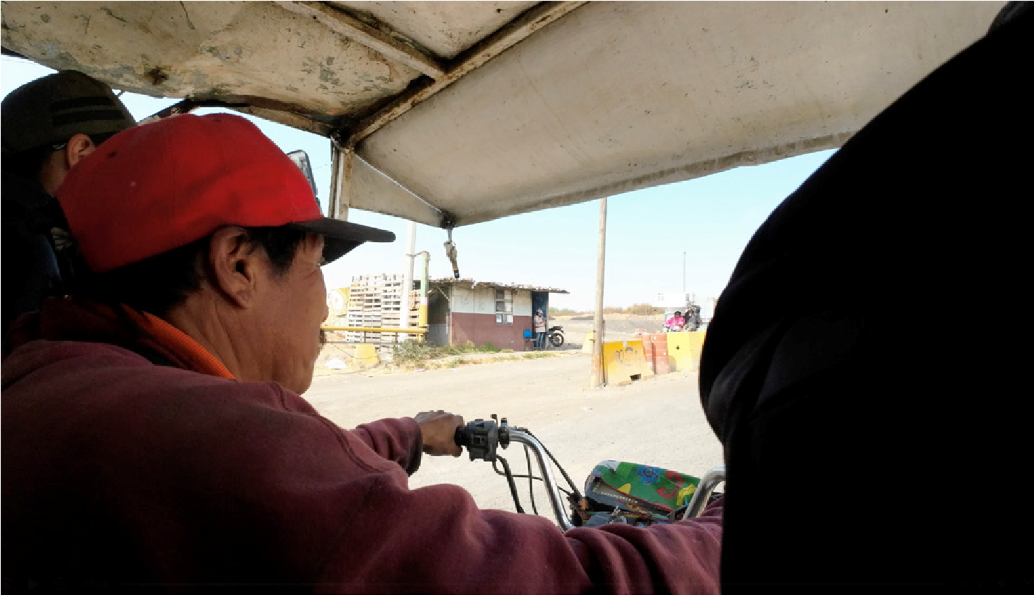 The shot shows Cirilio from inside the moto-cart, placing the viewer in the collector&rsquo;s position: we cross the entrance with him, we see what he sees. Cirilio drives and looks straight ahead. The guard is posted at the entrance and watches from the corner of his eye, but Cirilio avoids his gaze. This immersive framing reveals a form of strategic care: making no waves, staying discreet, crossing quickly to protect his load.