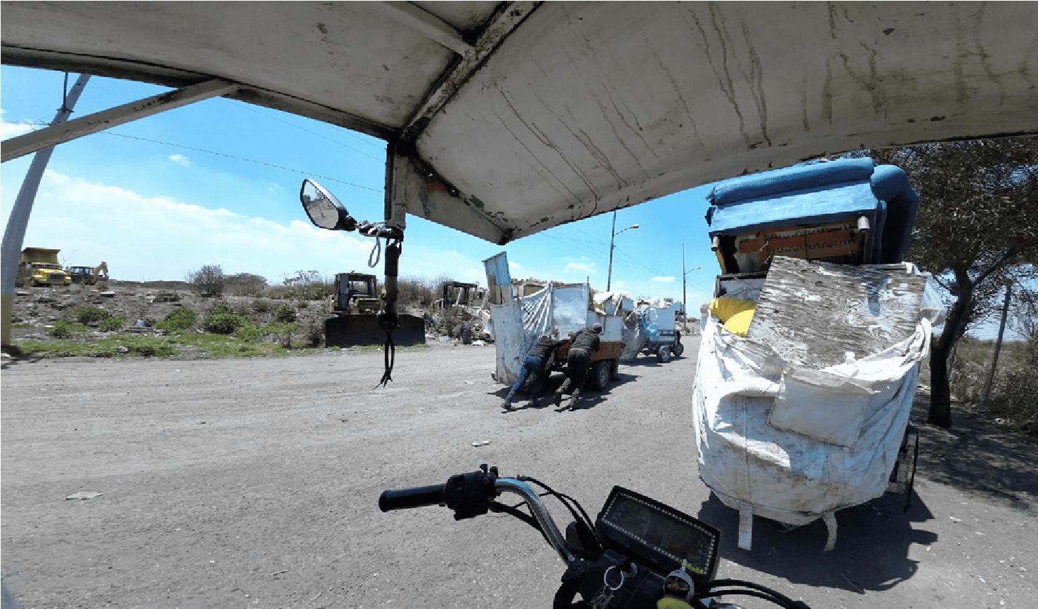 The wide angle from the handlebars of the moto-cart shows an everyday scene behind the entrance barrier: two collectors, among them Ana, pushing a moto-trailer. They are at the centre, surrounded by other moto-carts loaded with waste. The camera keeps its distance but films the scene unfolding: solidarity among collectors. It captures this spontaneous, collective, and immediate movement, without staging.