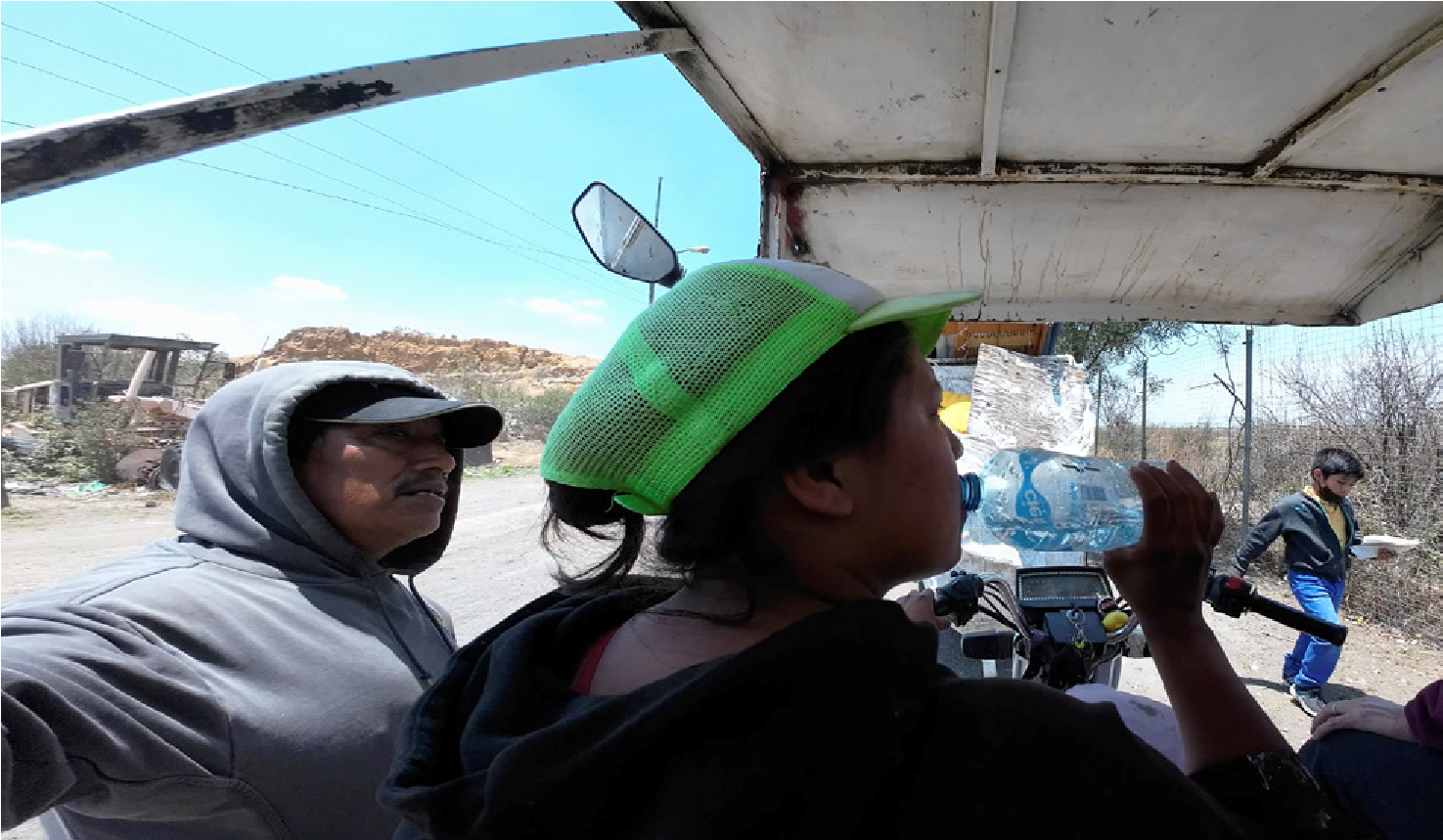 The framing is again intimate. It shows the interior of the vehicle and focuses on Ana&rsquo;s face as she drinks water offered to her in thanks for her help, while her father, tired from the effort, waits for his daughter to drink before hydrating himself. Off-frame, a glimpse of the landfill and its edge can be perceived to the right; heat and distant sounds are sensed. Care, understood as a moment of kindness and sharing, is rendered visible through the tightening of this scene.